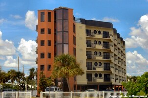 A multi-story hotel with balconies, a palm tree in front, and a clear sky.