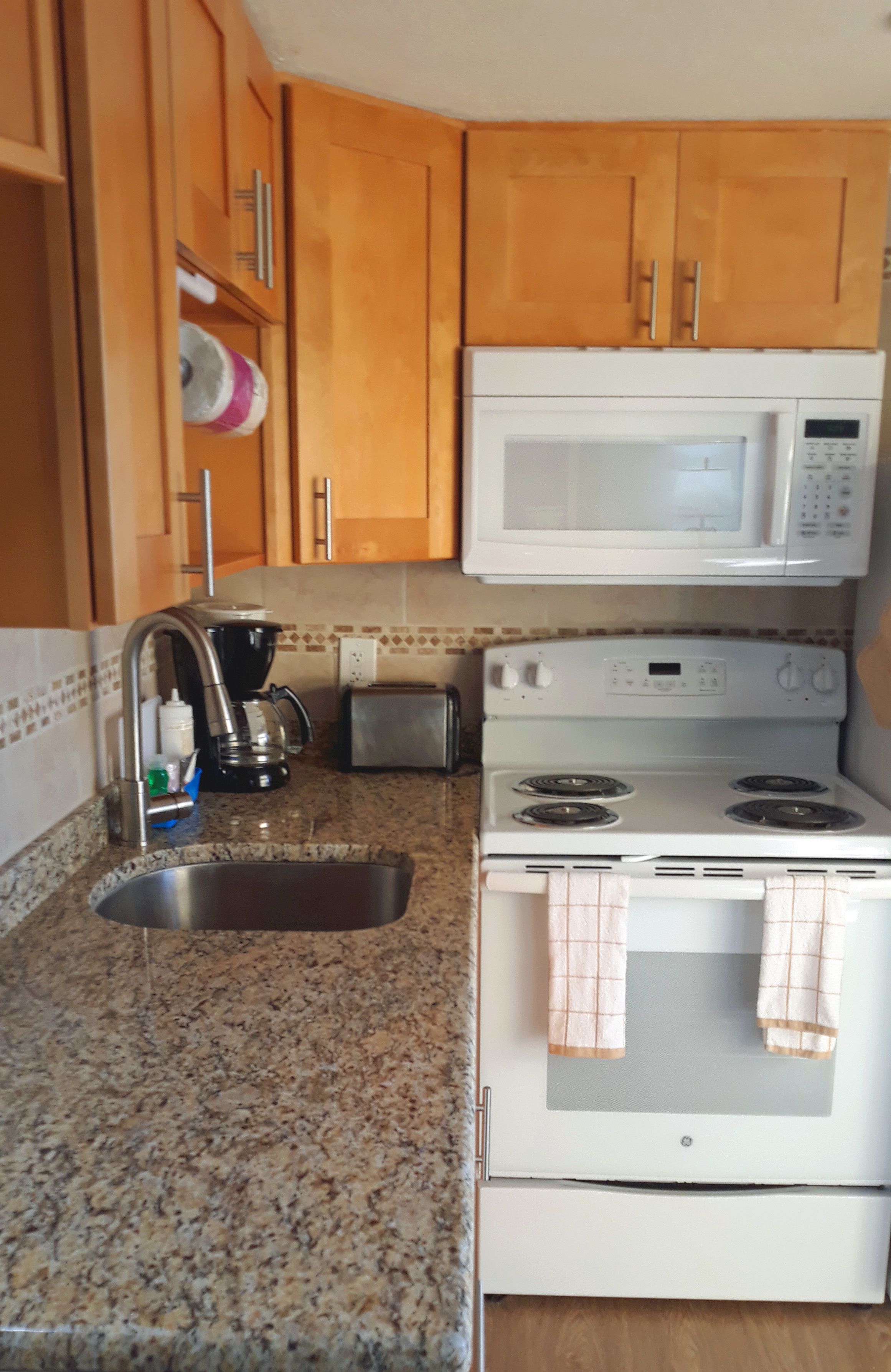 A compact kitchen with wooden cabinets, a microwave, a stove, a coffee maker, and a sink.