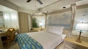 A hotel room with a bed, a desk, a chair, a nightstand, a lamp, a ceiling fan, and a mural of a beach scene.