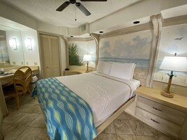 A hotel room with a bed, a desk, a chair, a nightstand, a lamp, a ceiling fan, and a mural of a beach scene.