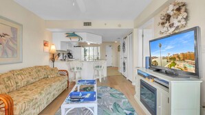 A living room with a sofa, a coffee table with remote controls, a TV, and a view of a pool and buildings.