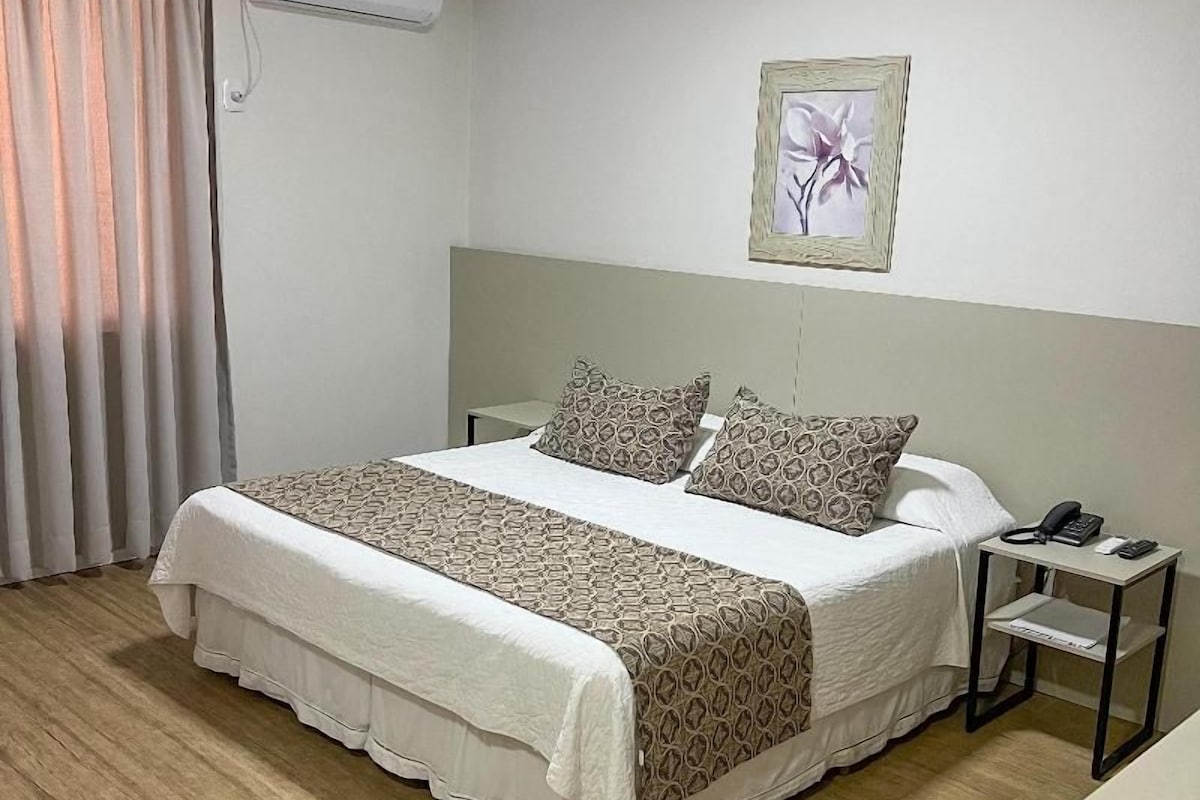 A hotel room with a bed, a bedside table with a phone, an air conditioning unit, and a window with curtains.