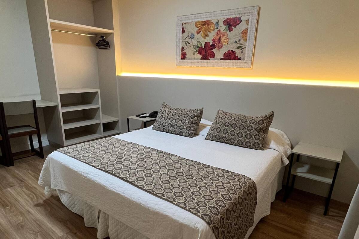 A neatly arranged bedroom with a large bed, a bedside table, a nightstand, a wardrobe, and a hanging shelf.