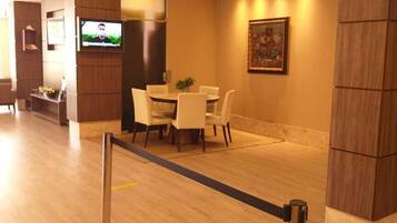 A hotel lobby with a wooden floor, a dining area with a table and chairs, a television mounted on the wall, and a framed picture on the wall.