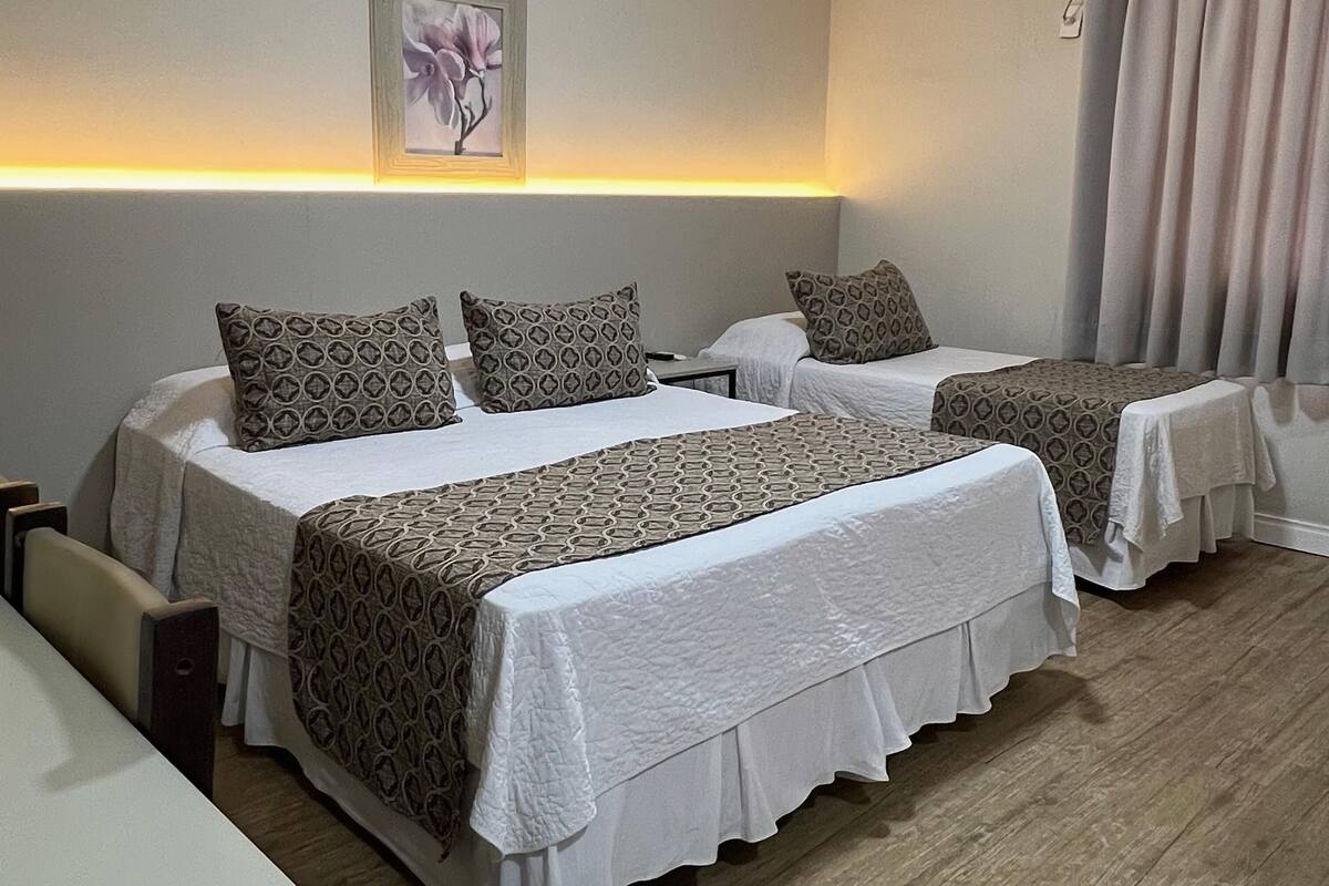 A hotel room with two beds, a bedside table, a chair, a wall-mounted air conditioner, and a window with curtains.