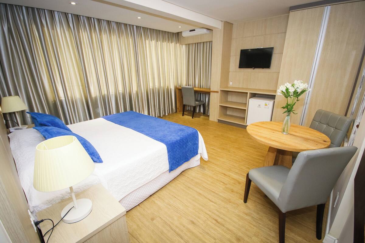 A hotel room with a bed, desk, chair, TV, and a small table with flowers.