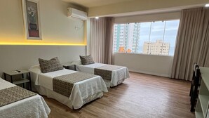 A hotel room with two beds, a bedside table, an air conditioning unit, a window with curtains, and a view of buildings outside.