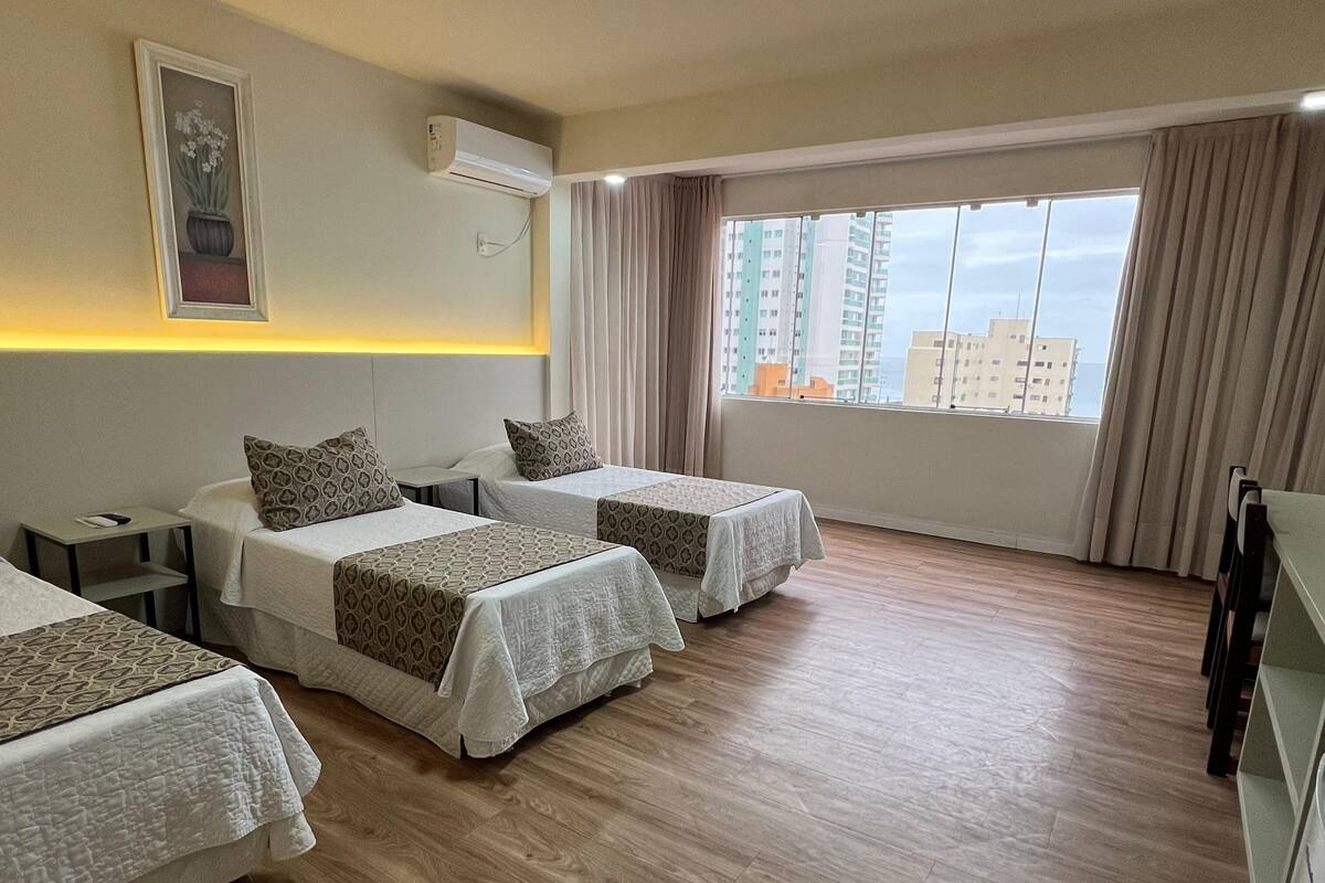 A hotel room with two beds, a bedside table, an air conditioning unit, a window with curtains, and a view of buildings outside.