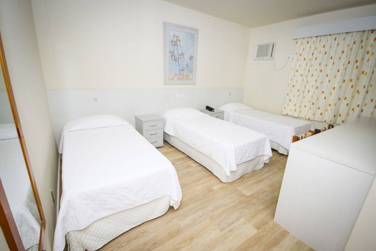 A hotel room with two single beds, a small bedside table with a lamp, a window with curtains, and a wall-mounted air conditioning unit.