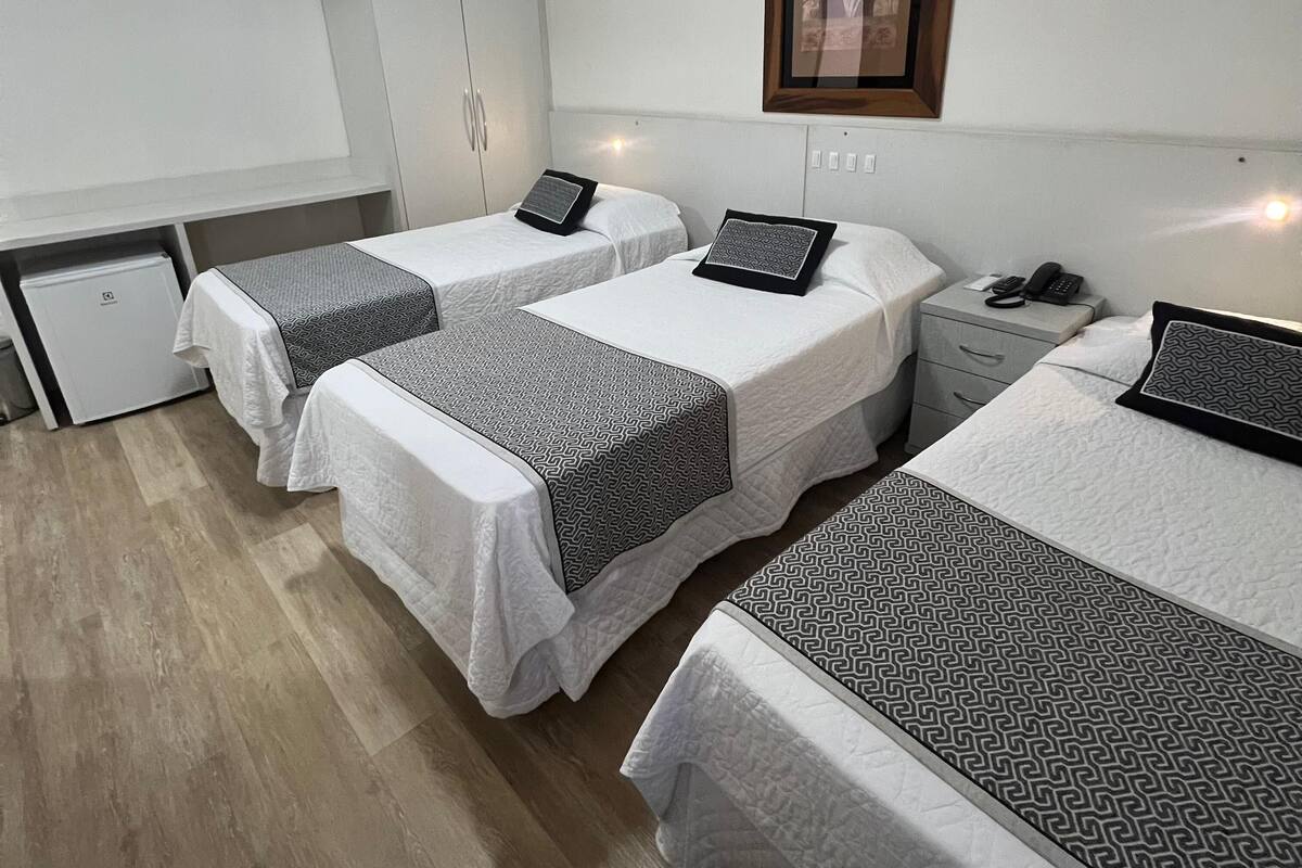 A hotel room with three beds, each with a patterned bedspread and pillows. There is a small desk with a telephone and a framed picture on the wall.