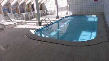 An indoor swimming pool with a tiled floor, surrounded by white lounge chairs and a covered seating area with a view of the sky.