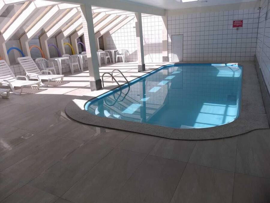 An indoor swimming pool with a tiled floor, surrounded by white lounge chairs and a covered seating area with a view of the sky.