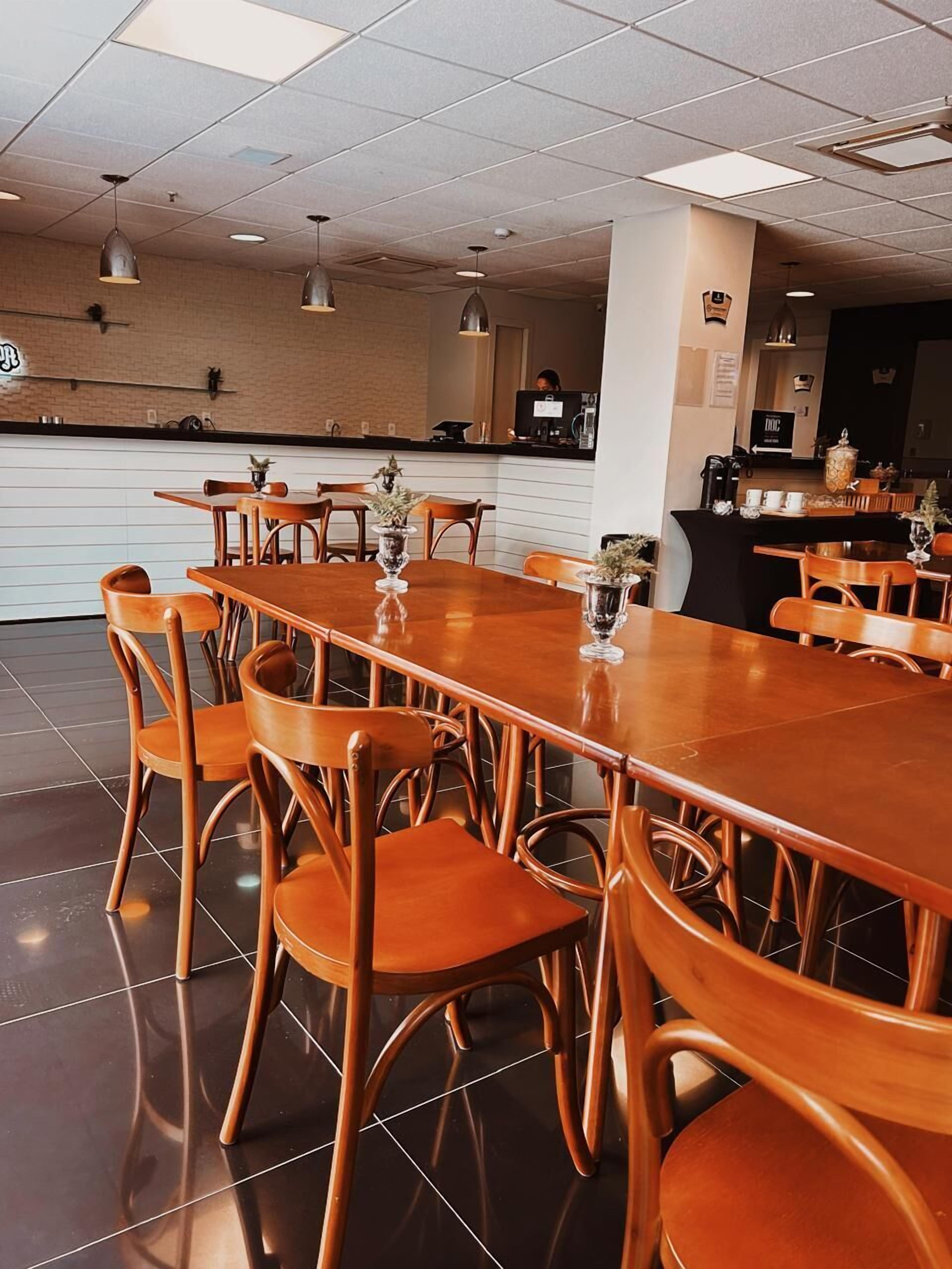 Uma sala de jantar ampla, com uma longa mesa de madeira e cadeiras combinando, um balcão com máquina de café e um chão limpo, revestido de azulejos.