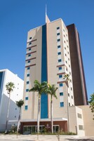 Un hotel alto con una banda vertical azul en el centro, varios pisos y palmeras en primer plano.