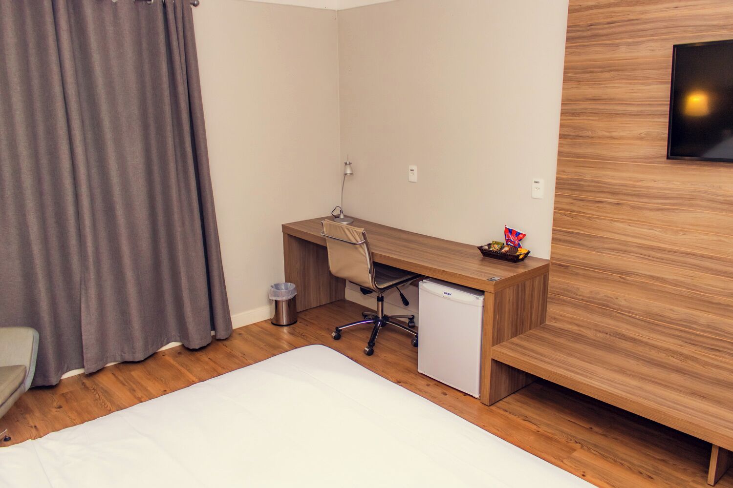 A hotel room with a wooden desk, a chair, a small refrigerator, a trash bin, and a bed.