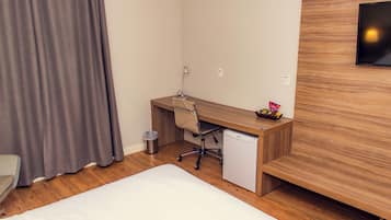 A hotel room with a wooden desk, a chair, a small refrigerator, a trash bin, and a bed.
