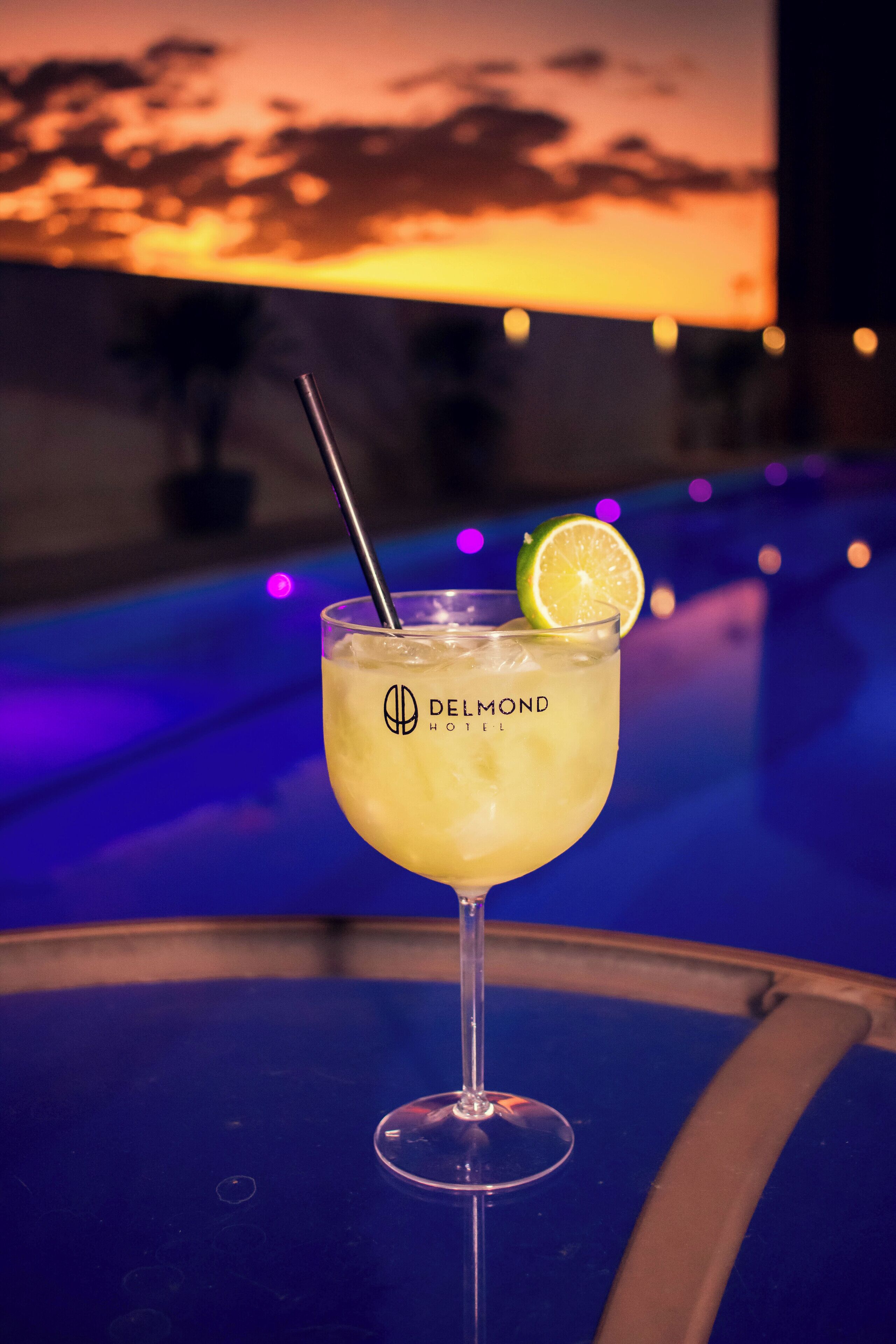 Un verre à cocktail, garni d’une tranche de citron vert, est posé sur une table d’où l’on peut admirer un coucher de soleil.