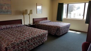 A hotel room with two beds, a nightstand, a lamp, a window with curtains, and a painting on the wall. - Safari Inn (Winston)