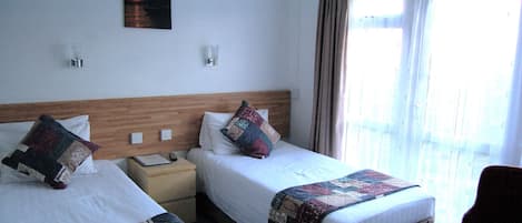 Een hotelkamer met twee bedden, een houten hoofdbord, een nachtkastje, een lamp en een raam met gordijnen.