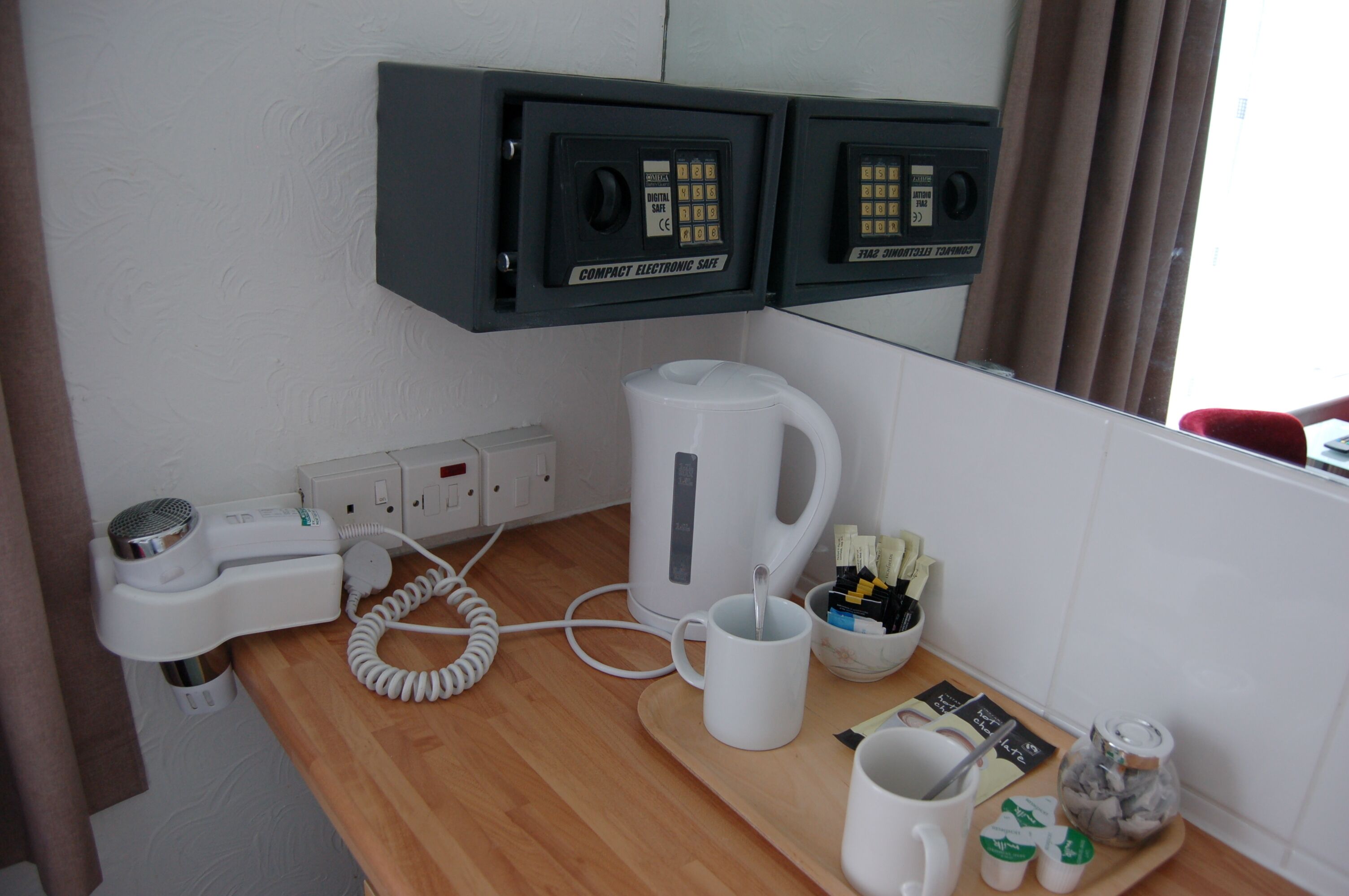 A compact electronic safe, a hairdryer, a kettle, and a coffee maker on a wooden counter.