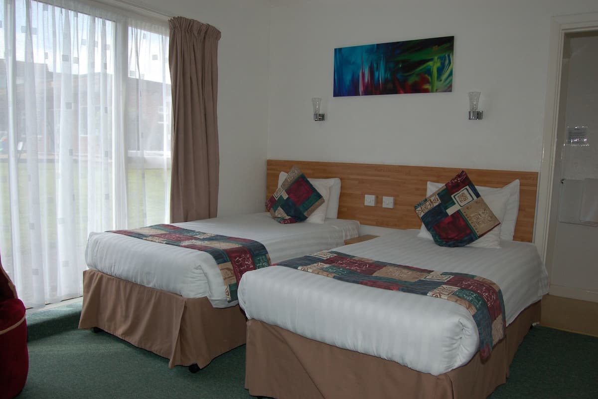 A hotel room with two beds, a large window with curtains, a painting on the wall, and a door.