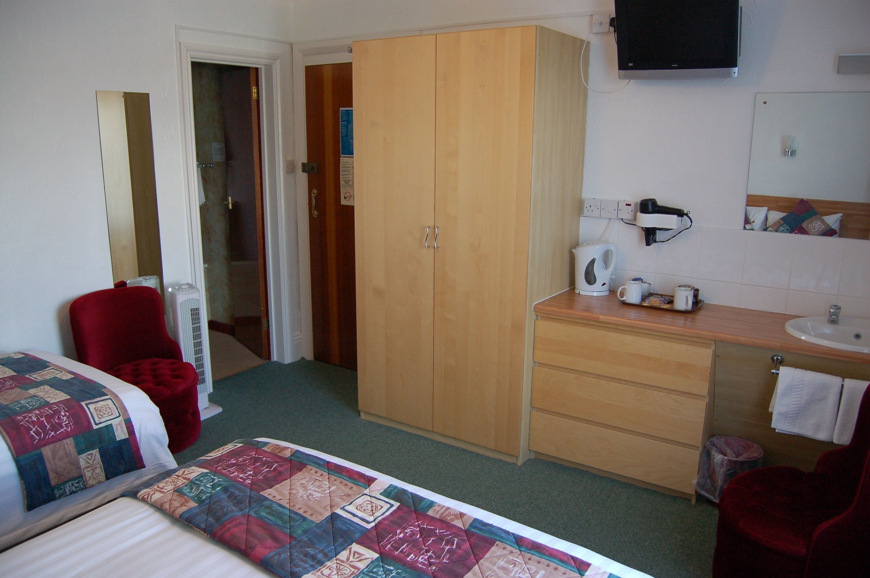A hotel room with a bed, a red chair, a wooden wardrobe, a bathroom with a sink and mirror, and a television on the wall.