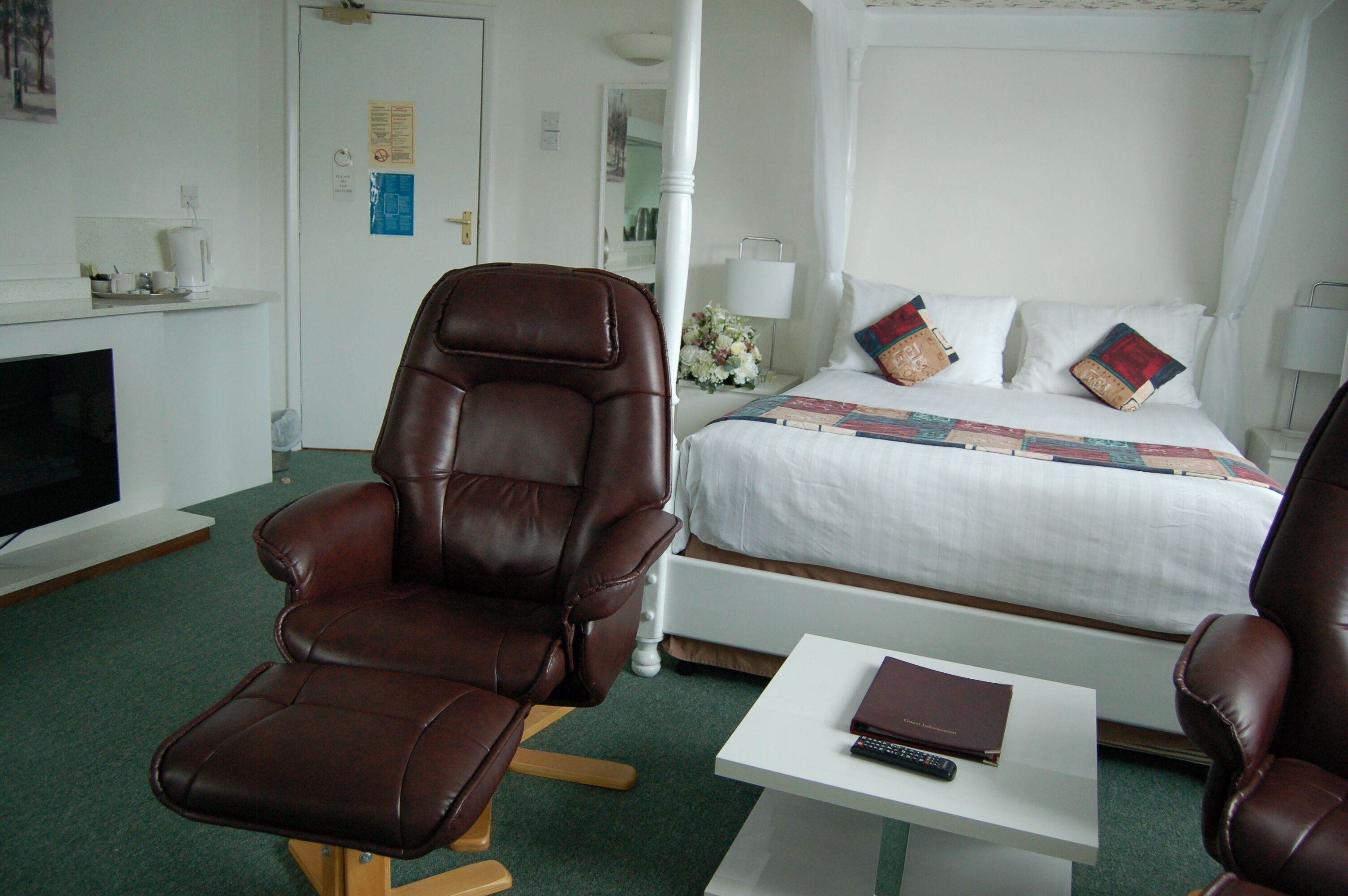 Una camera d'albergo con un letto grande, una poltrona in pelle marrone, un comodino bianco con un telecomando e una televisione su un mobile.