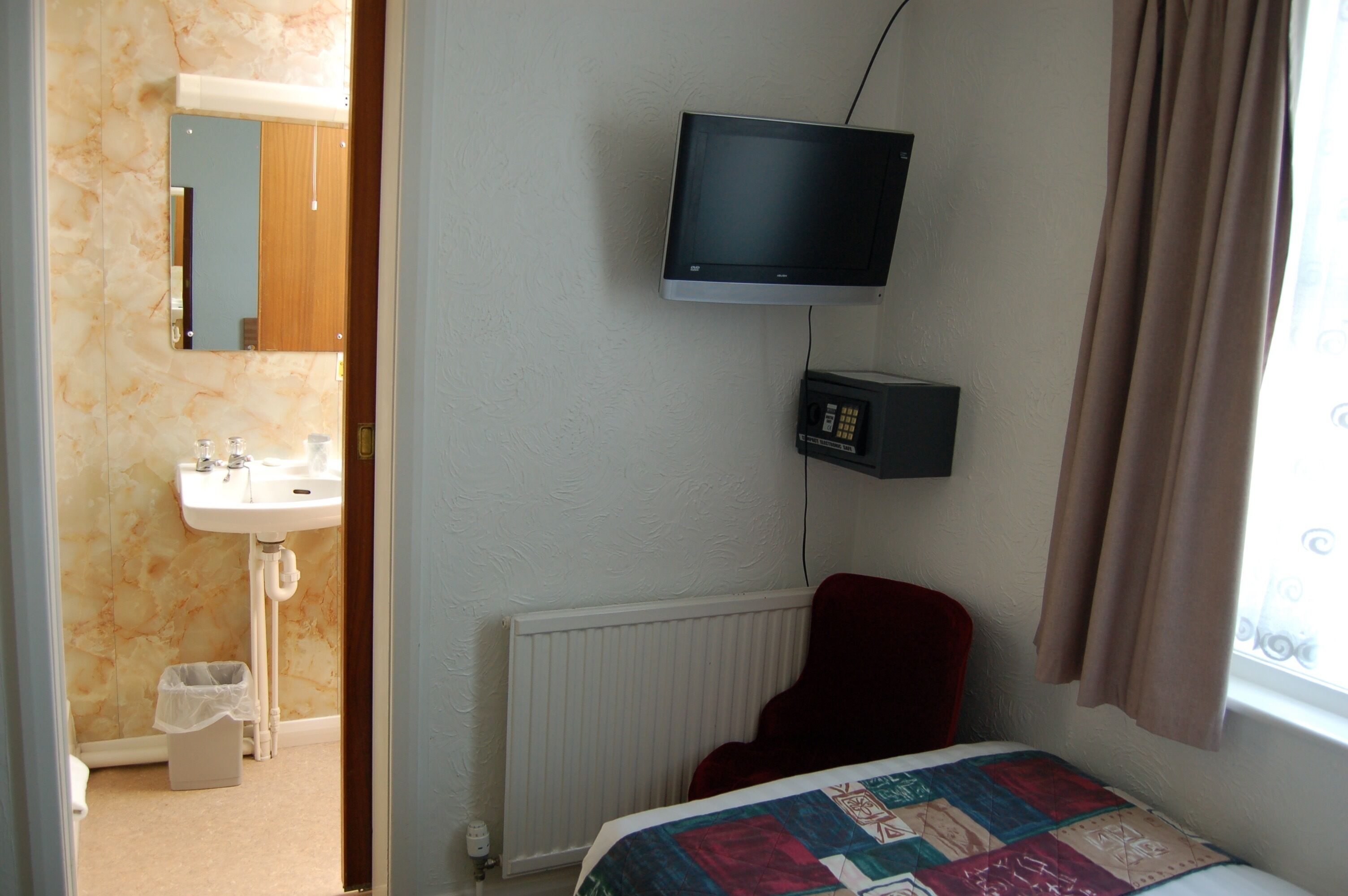 A hotel room with a bed, a television mounted on the wall, a red chair, a small window with curtains, a radiator, and a bathroom with a sink and mirror.