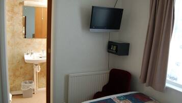 A hotel room with a bed, a television mounted on the wall, a red chair, a small window with curtains, a radiator, and a bathroom with a sink and mirror.