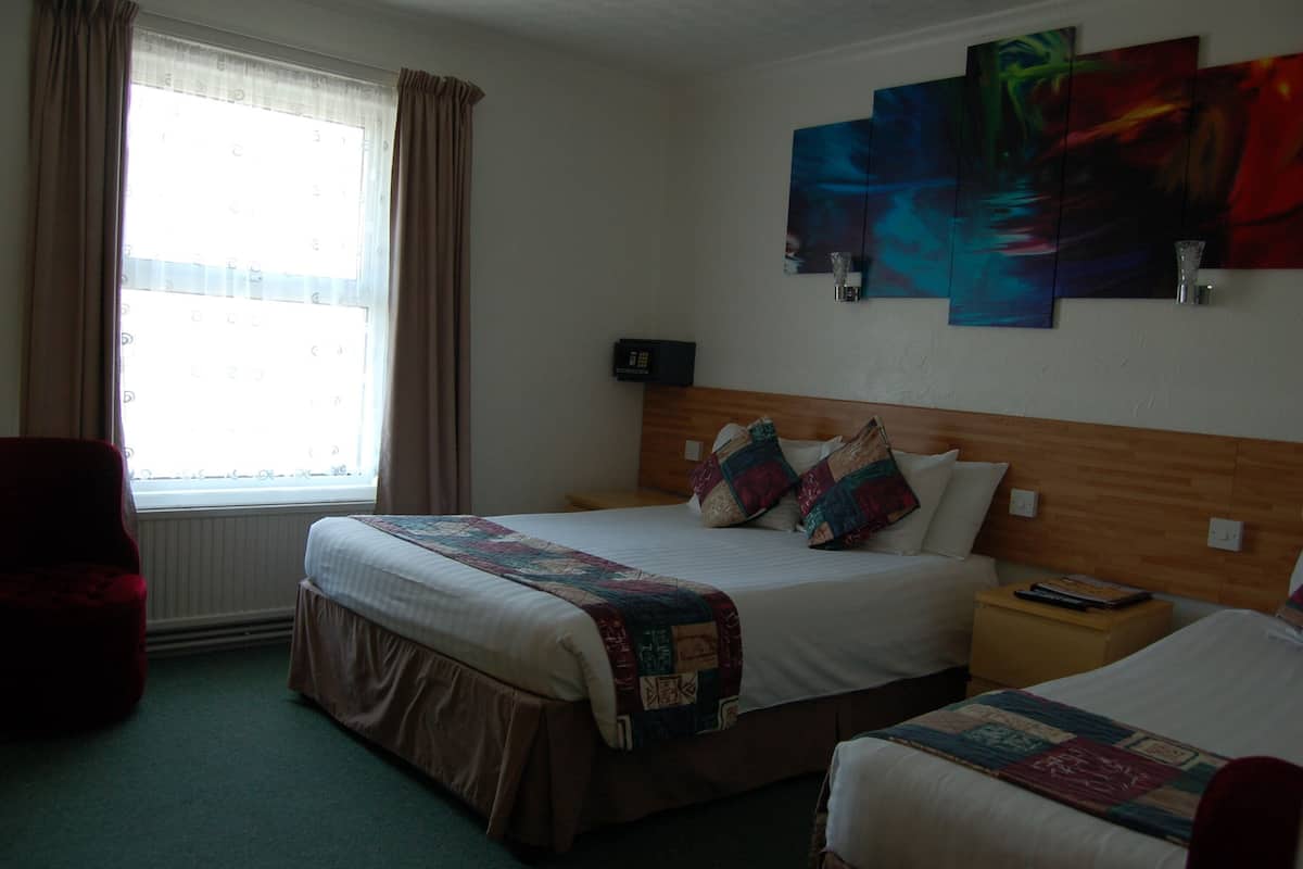 A hotel room with a large bed, a smaller bed, a nightstand, a chair, a window with curtains, and abstract wall art.