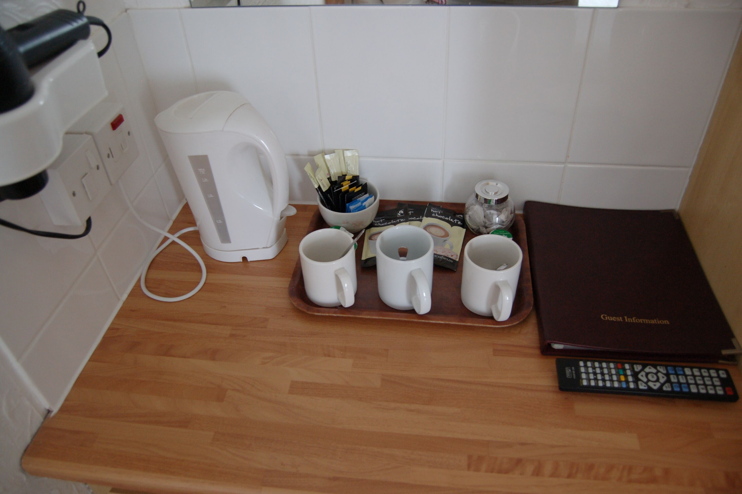 A kitchen counter with a coffee maker, a tray with four mugs, a remote control, and a guest information booklet.