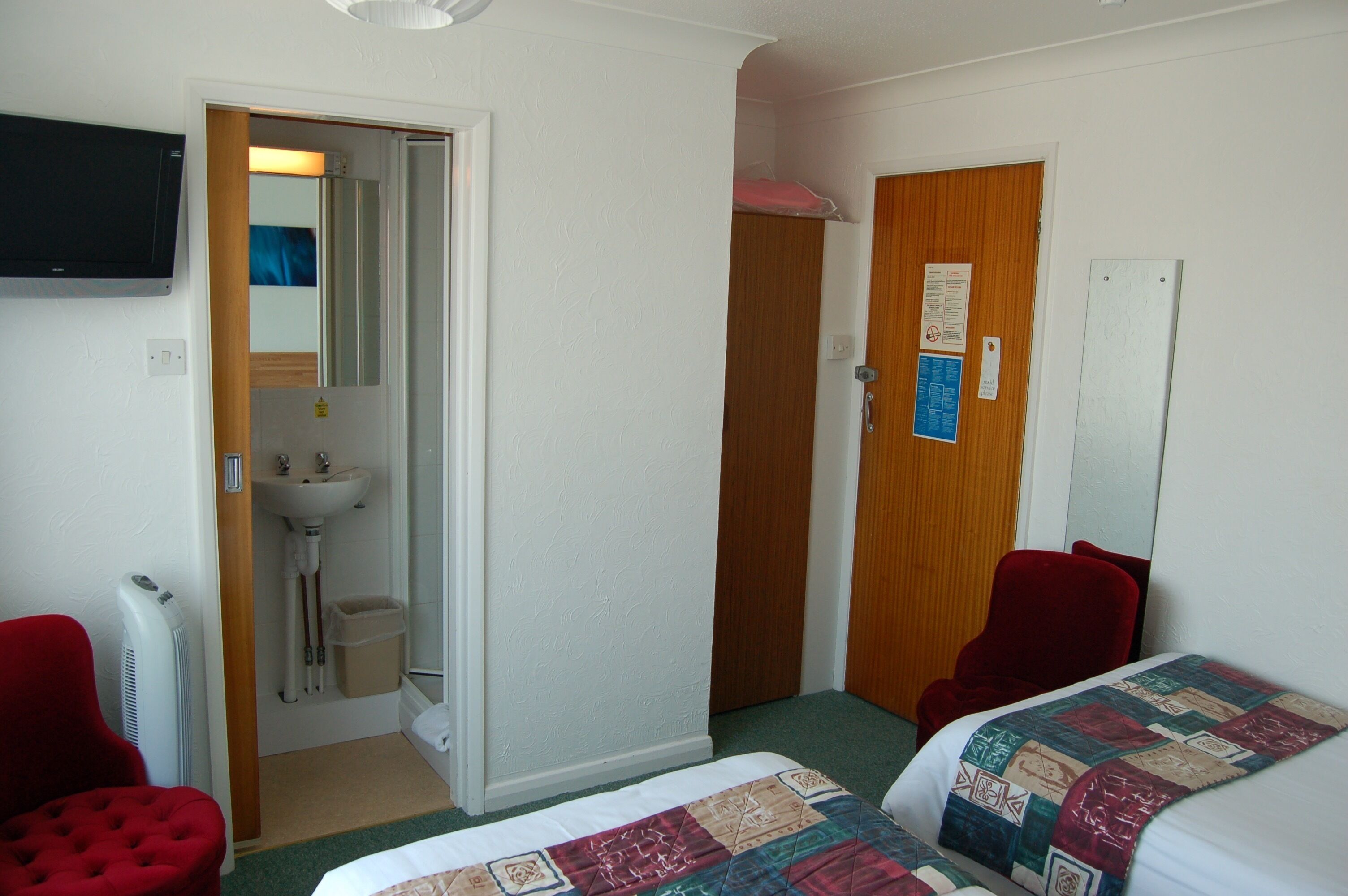 Une chambre d’hôtel avec deux lits, une chaise rouge, une porte en bois, un lavabo blanc et un miroir.