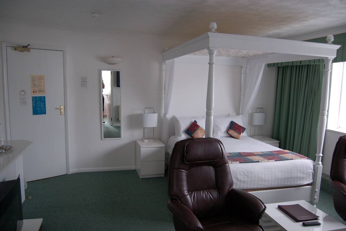A four-poster bed with a canopy, a nightstand, a mirror, and a chair in a hotel room.