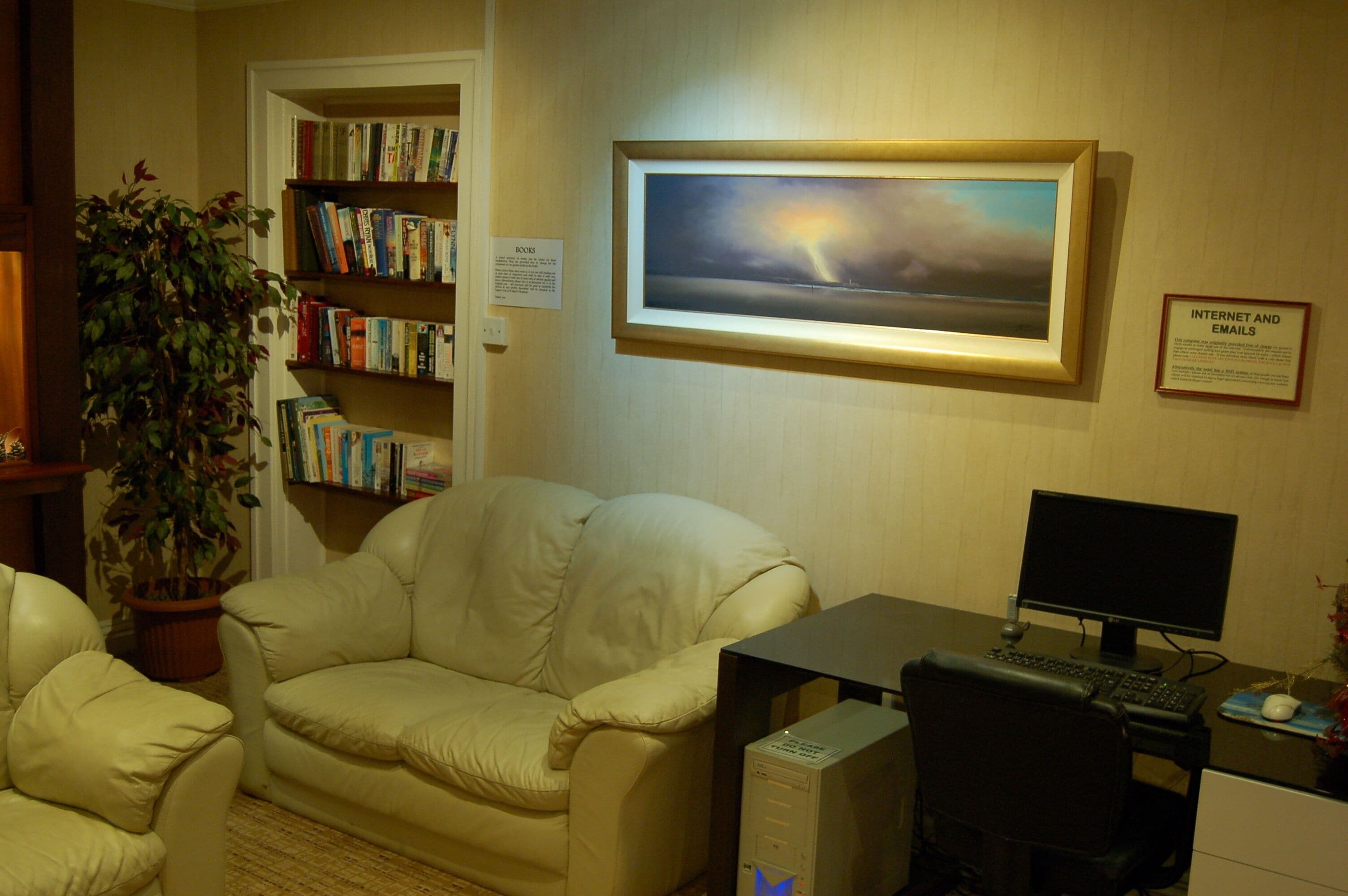 A room with a beige sofa, a bookshelf, a framed painting, a desk with a computer, and a potted plant.