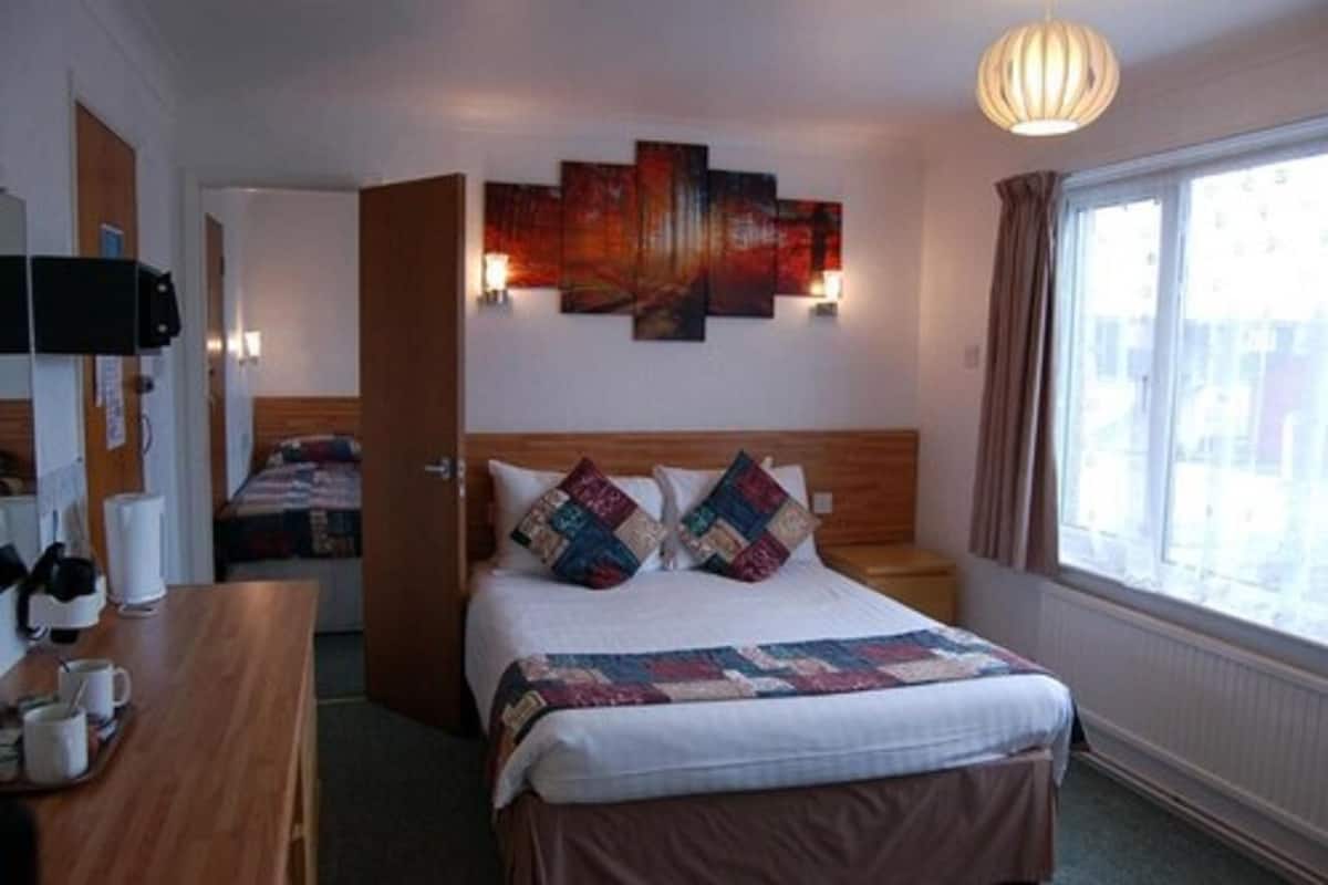 A hotel room with a bed, bedside table, lamp, artwork, and a window with curtains.