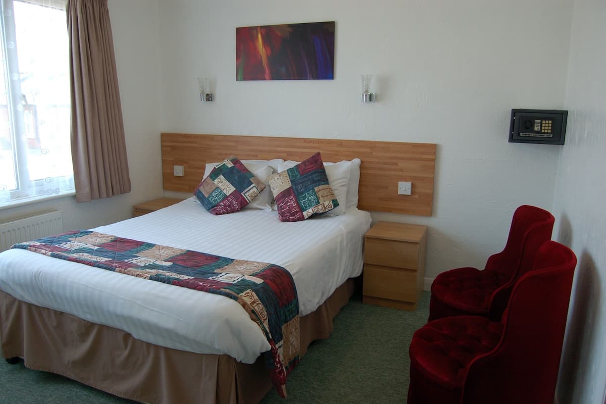 A neatly made bed with a quilt, two bedside tables, a wall-mounted safe, and a red chair.