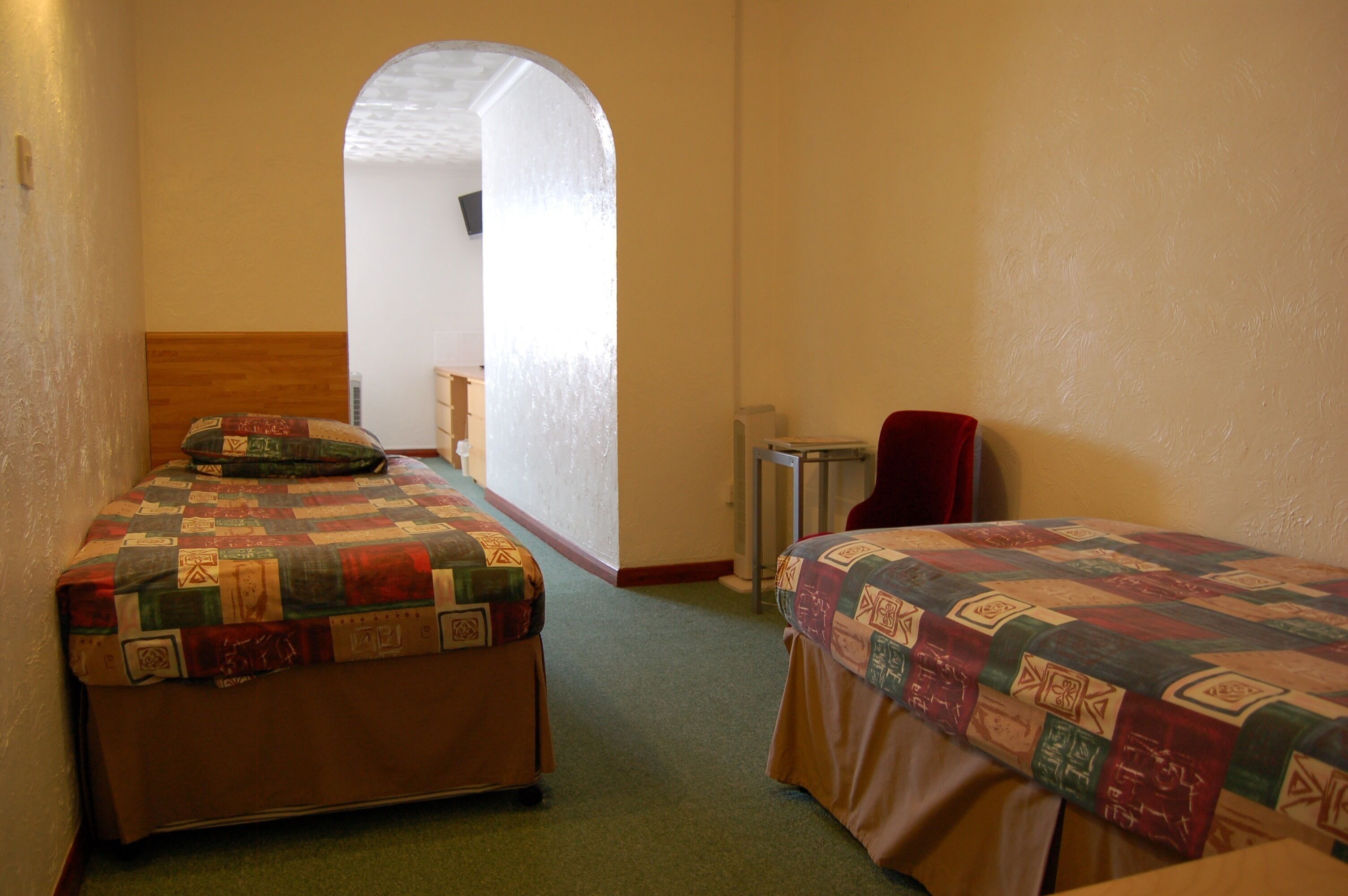 A room with two beds, each with a quilted bedspread, a wooden headboard, and a red chair.