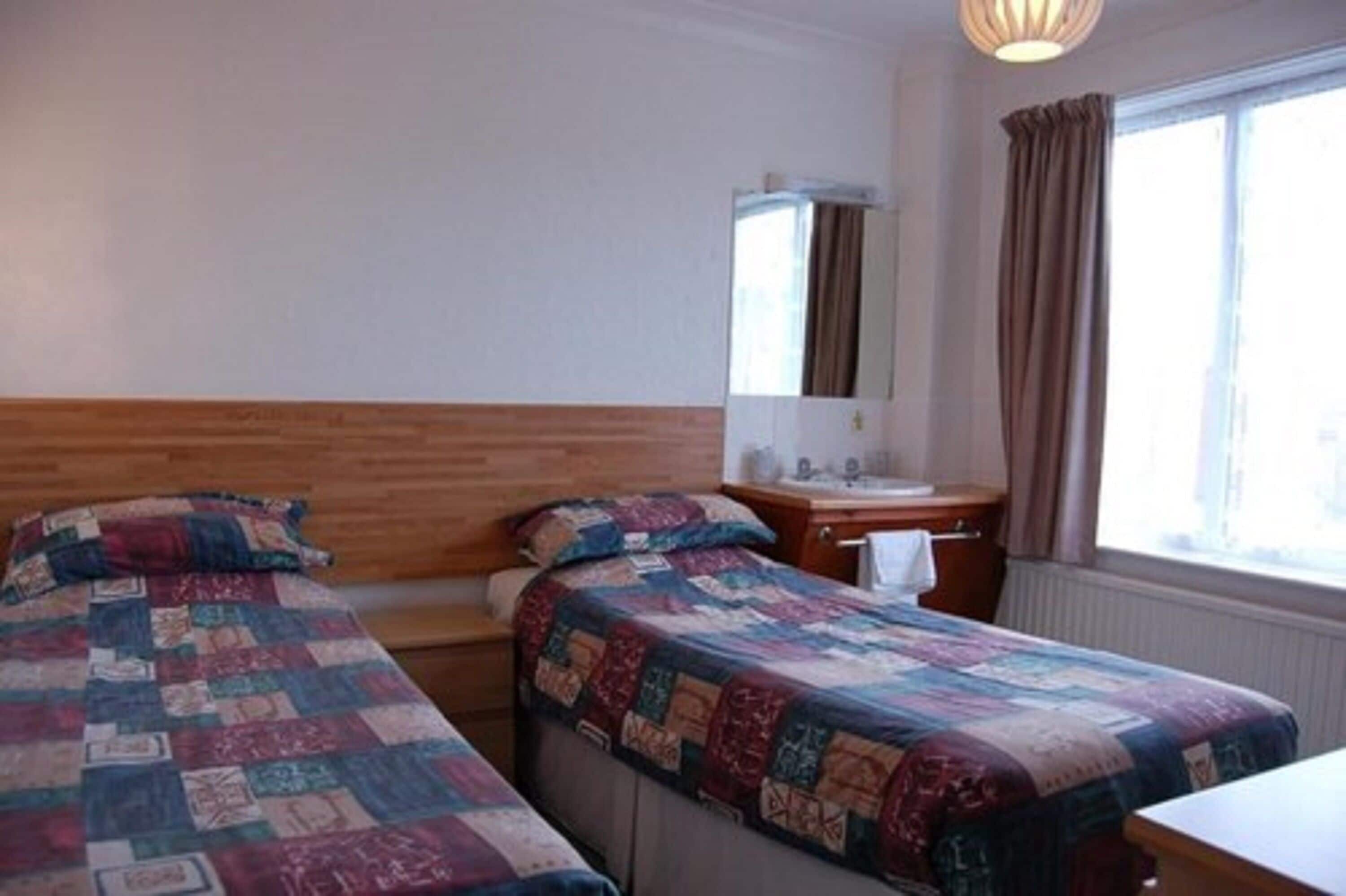A room with two beds, a wooden headboard, a small bedside table with a mirror, a window with curtains, and a radiator.
