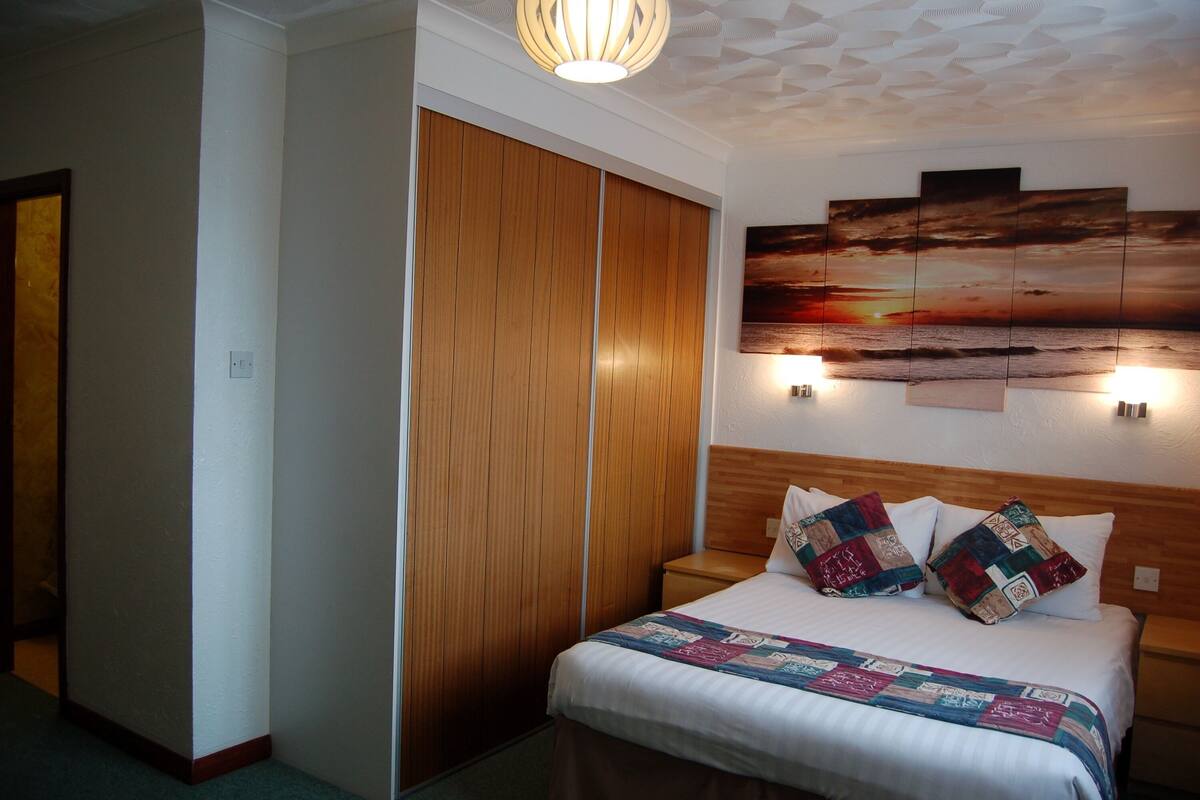 A bedroom with a wooden wardrobe, a bed with a quilt, and a sunset painting on the wall.