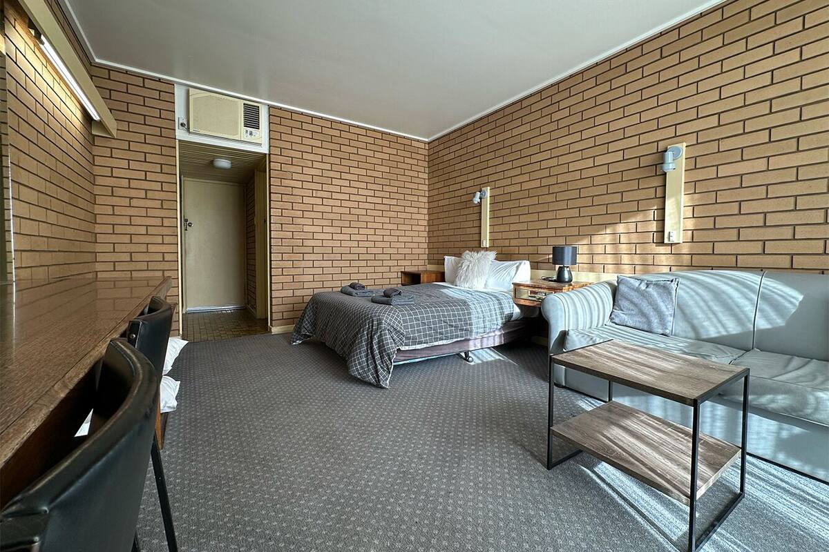 Una habitación de hotel con pared de ladrillo, una cama con colcha a cuadros, un sofá, una mesita y un escritorio con una silla.