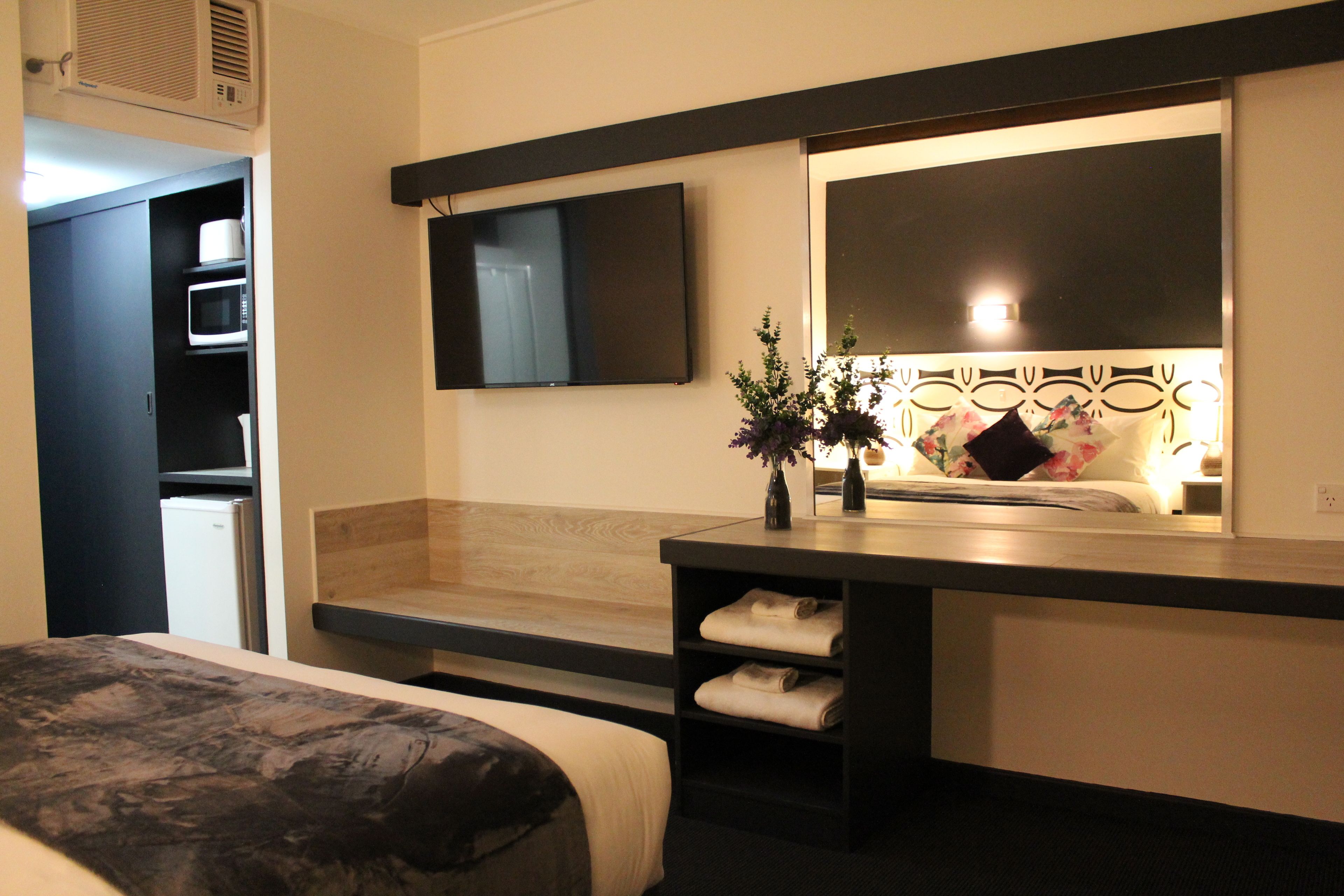 A hotel room with a bed, a TV mounted on the wall, a small table with a vase, and a wooden bench.