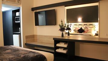 A hotel room with a bed, a TV mounted on the wall, a small table with a vase, and a wooden bench.