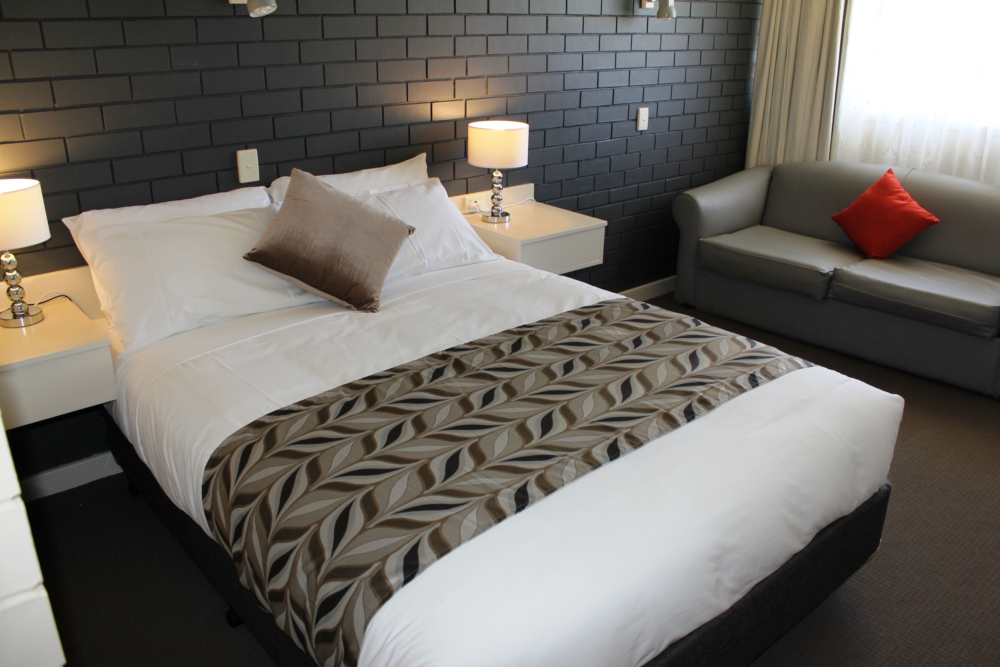 Una habitación de hotel con una cama, una mesita de noche con una lámpara, un sofá con un cojín rojo y una pared con diseño de ladrillo.