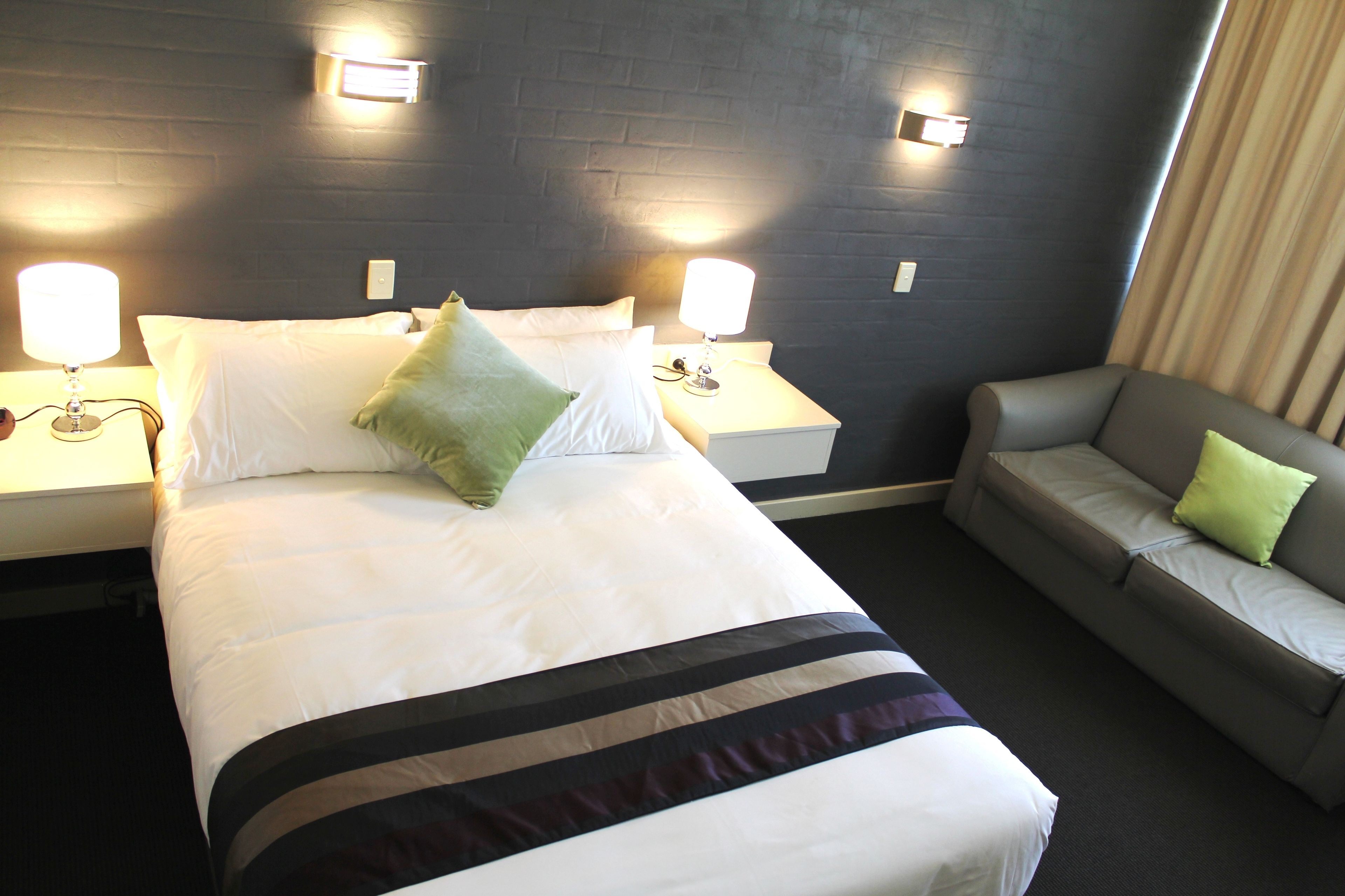 Una habitación de hotel con una cama, dos lámparas de noche, un sofá gris y un aplique de pared.