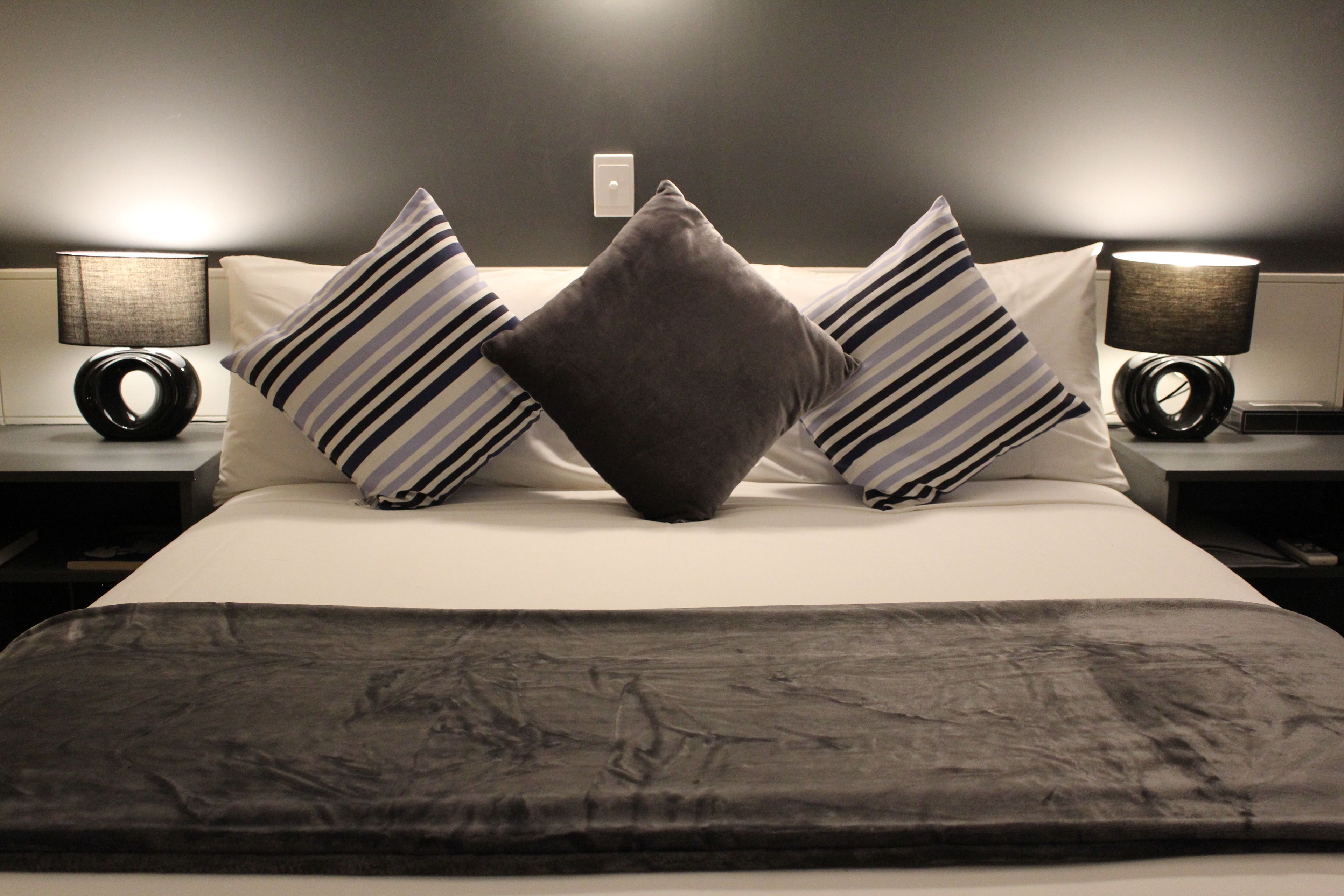 A neatly made bed with striped pillows and a dark throw blanket, flanked by two bedside tables with lamps.