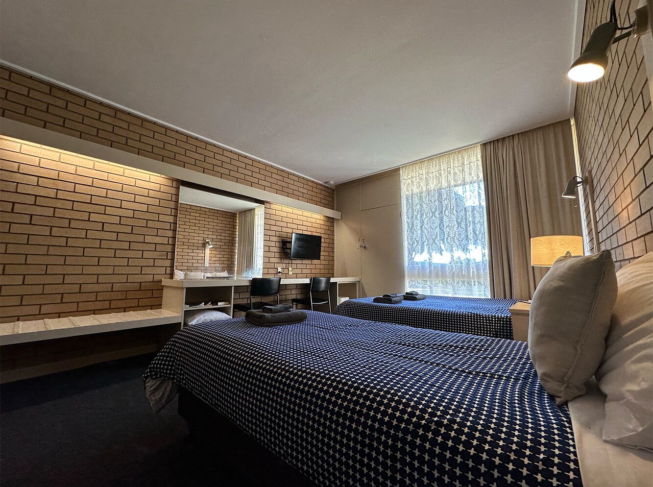 A hotel room with two beds, a desk, a chair, a TV, and a window with curtains.