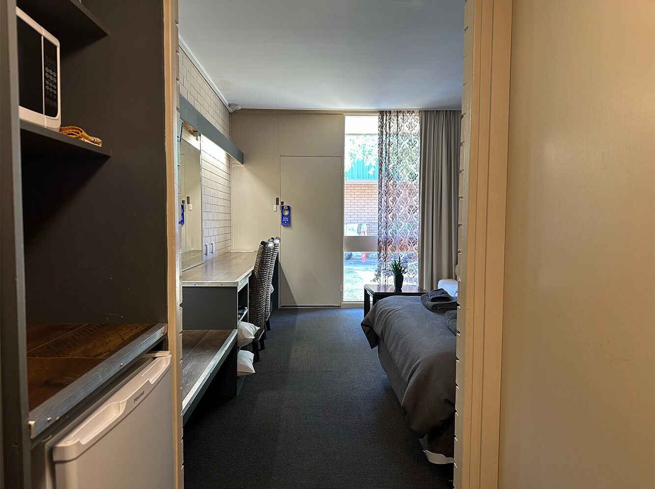 A compact hotel room with a kitchenette, a bed, a small table, and a window with curtains.