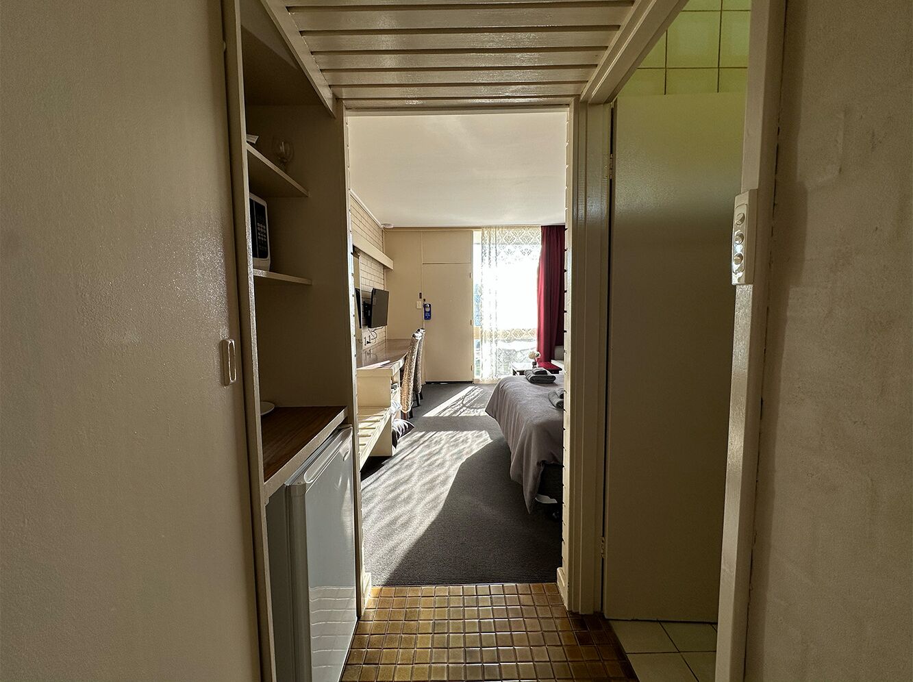 A narrow hallway leading to a bedroom with a bed, a small table, and a chair. There is a kitchenette area with a microwave and a sink.