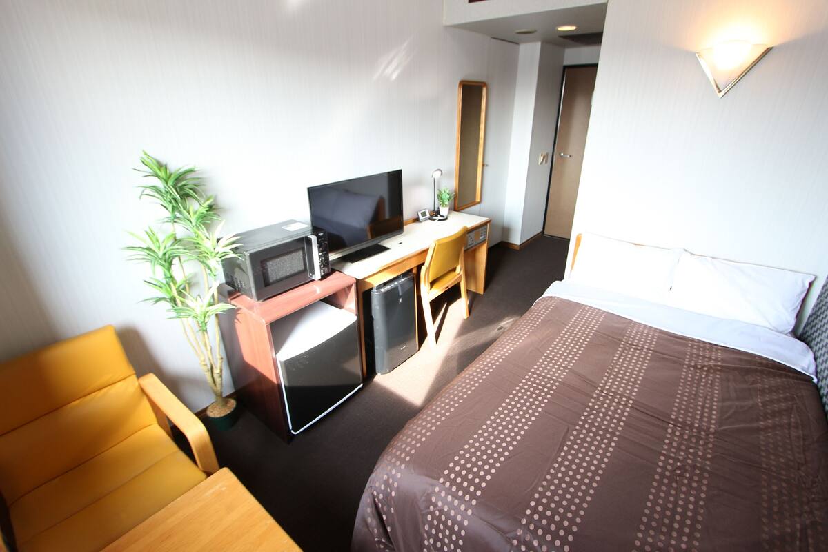 Habitación de hotel con cama, escritorio con computadora, silla, refrigerador pequeño y planta en maceta.