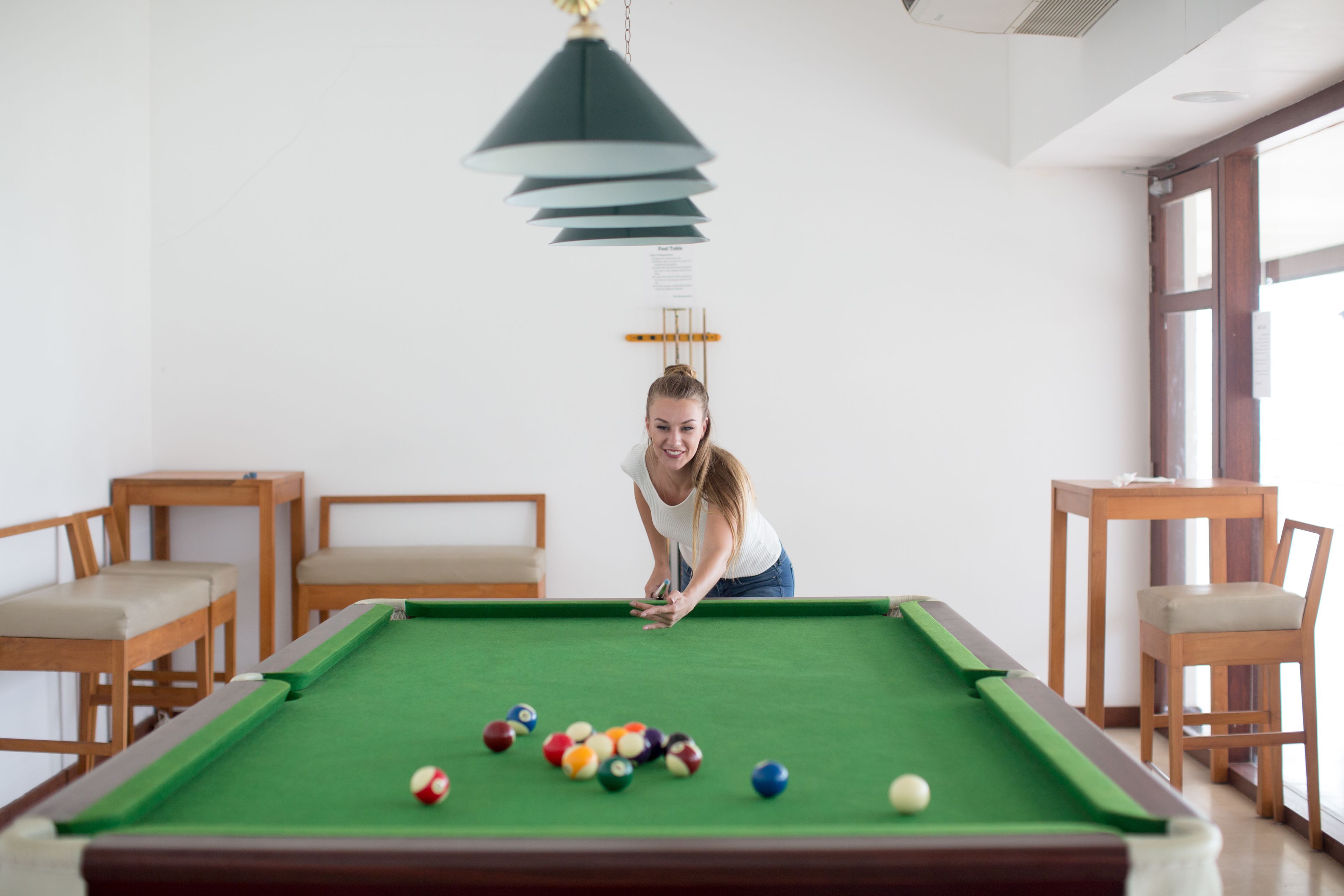 A pool table with balls, a hanging lamp, and wooden furniture in a room with white walls.